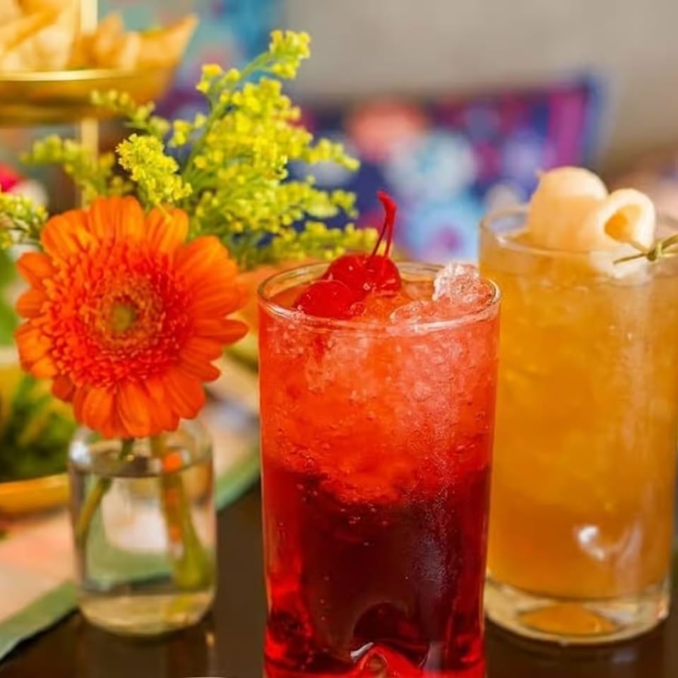 Two vibrant cocktails, one red with cherries and one amber with lychee, next to an orange flower.