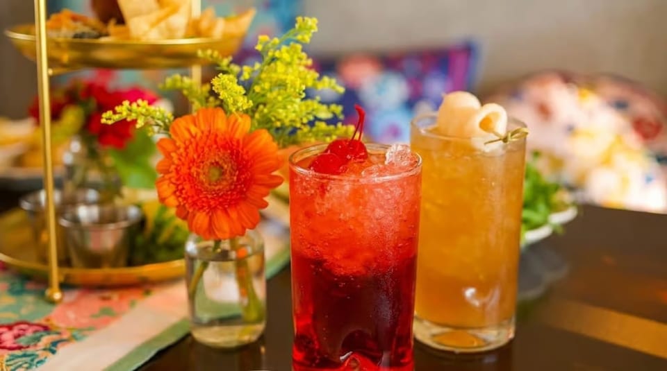 Two vibrant cocktails, one red with cherries and one amber with lychee, next to an orange flower.