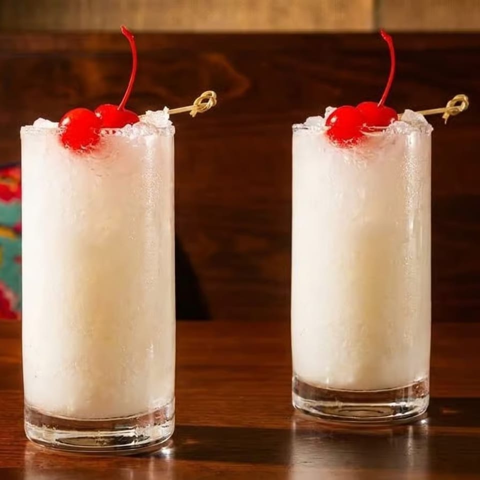 Two tall, frosty white cocktails with red cherries and crushed ice on a wooden table.