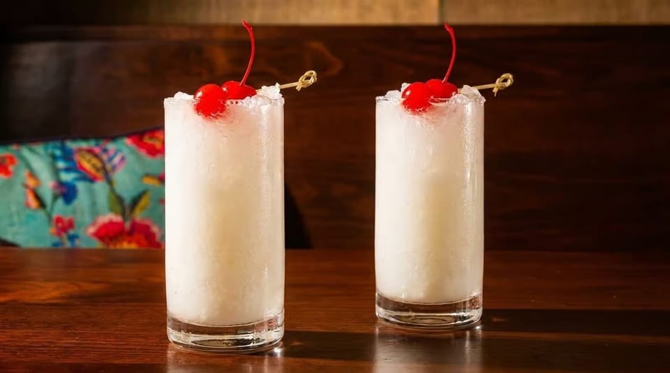 Two tall, frosty white cocktails with red cherries and crushed ice on a wooden table.