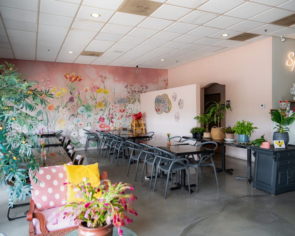 Restaurant interior with a floral mural, dark tables, gray chairs, and potted plants.