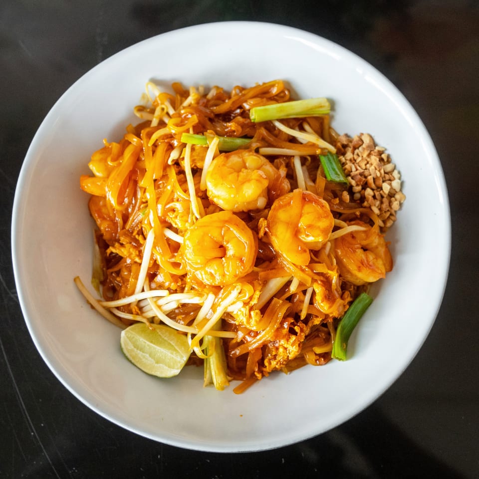 A white bowl of Pad Thai with shrimp, bean sprouts, green onions, peanuts, and a lime wedge.