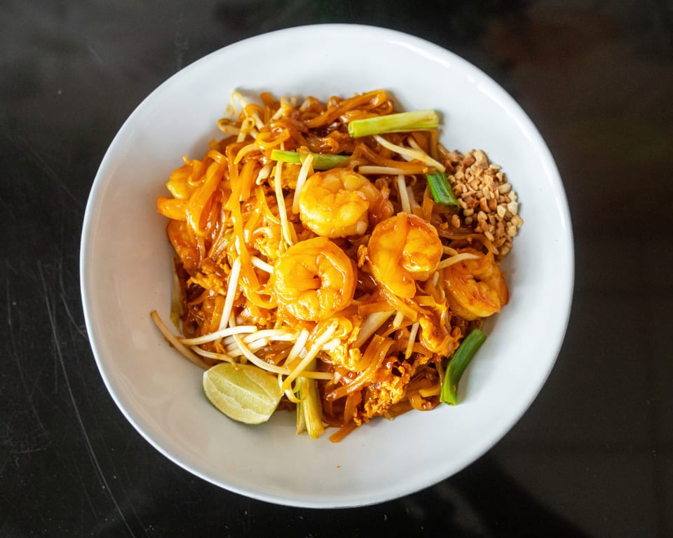 A white bowl of Pad Thai with shrimp, bean sprouts, green onions, peanuts, and a lime wedge.