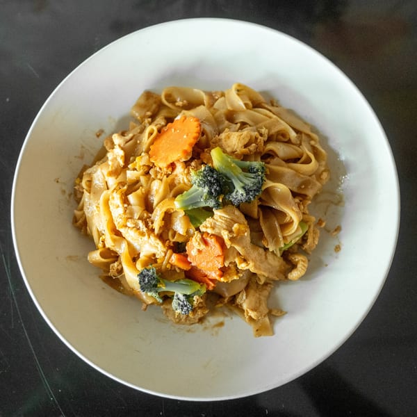 A white bowl of Pad See Ew with chicken, broccoli, and carrots on a dark surface.