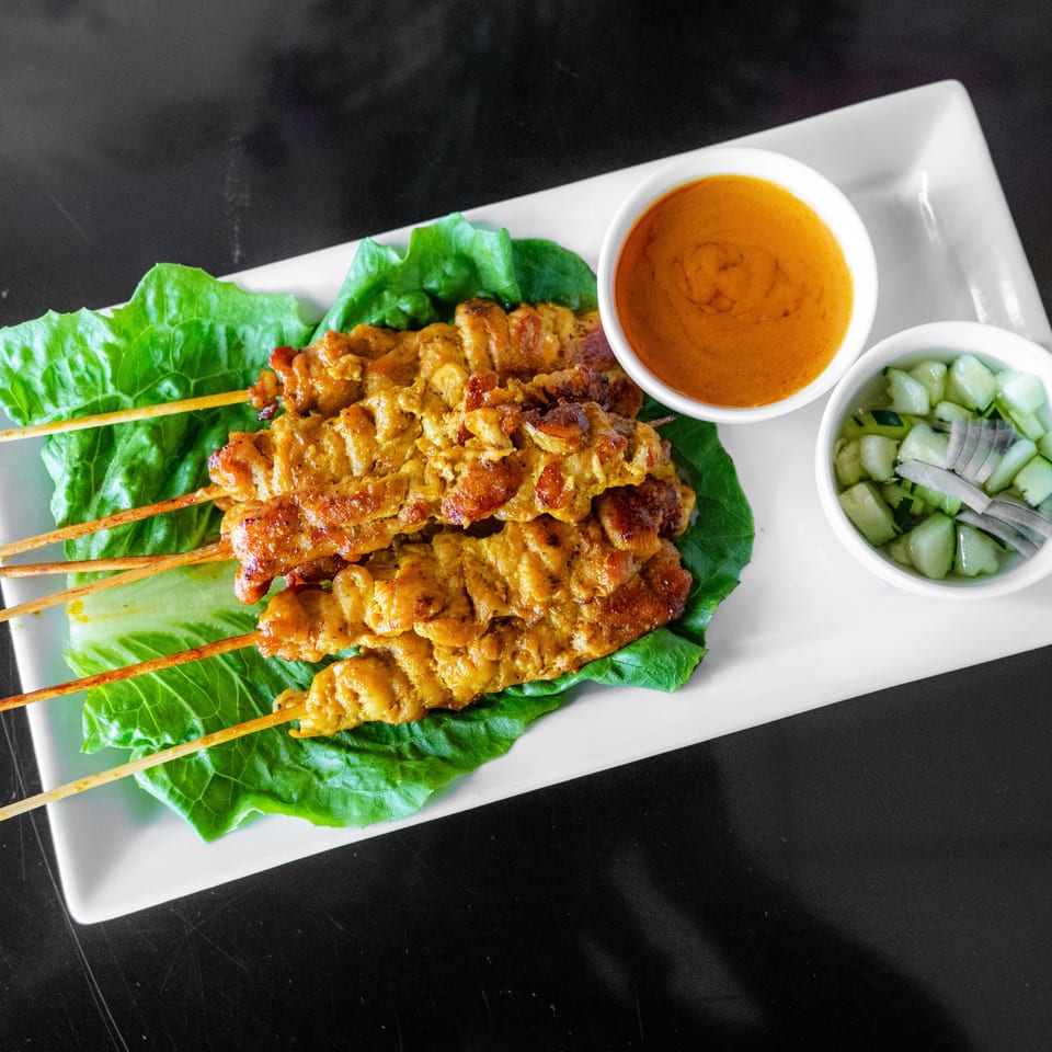 Chicken satay skewers on lettuce with peanut sauce and cucumber relish on a white plate.