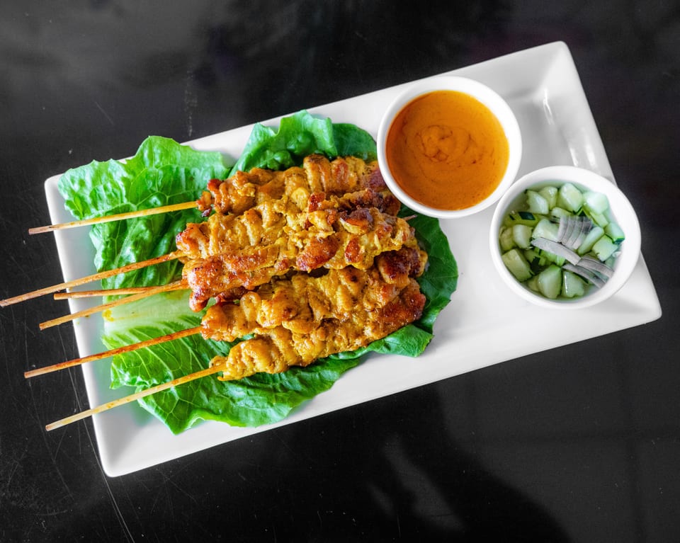 Chicken satay skewers on lettuce with peanut sauce and cucumber relish on a white plate.