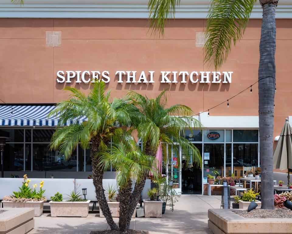 Spices Thai Kitchen storefront with outdoor seating, palm trees, and an "OPEN" sign in the window.