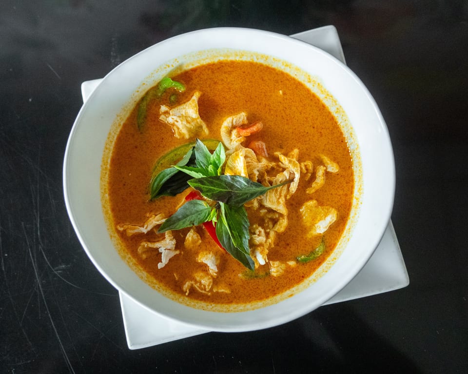 A white bowl of red curry with chicken, basil, and peppers on a dark surface.