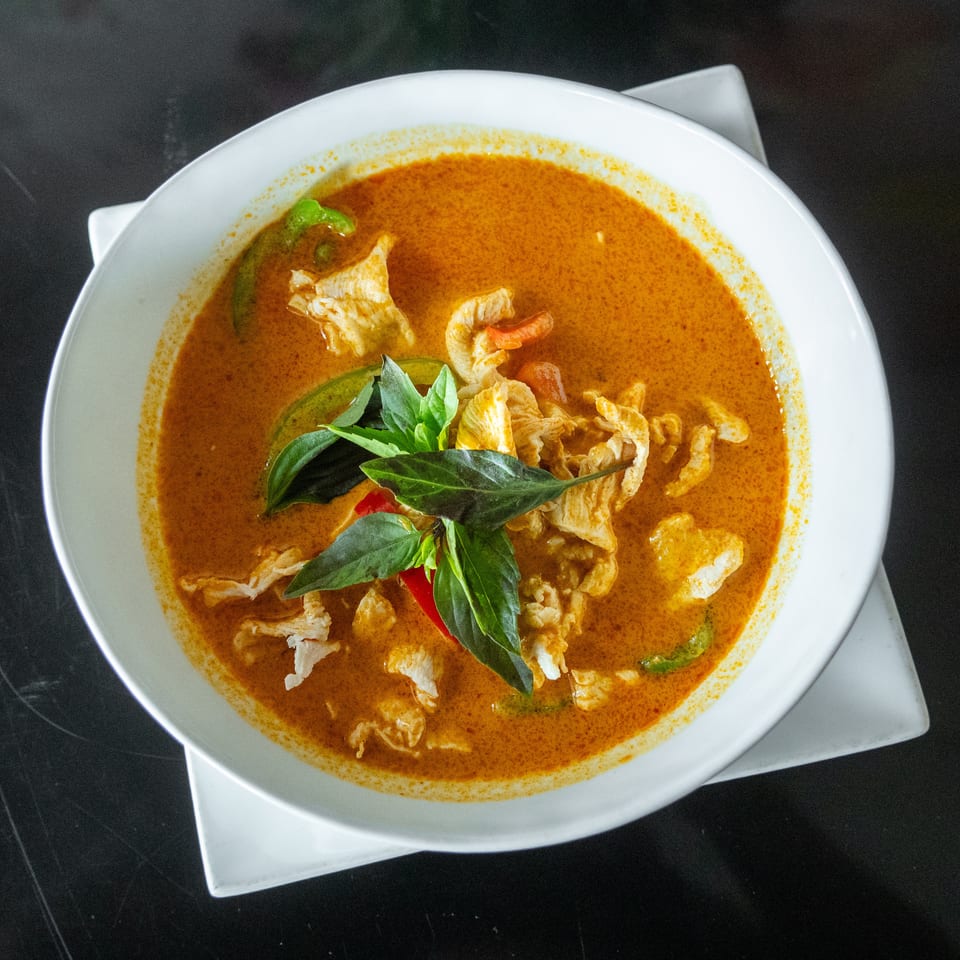 A white bowl filled with orange curry, chicken, green peppers, and basil leaves on a dark surface.