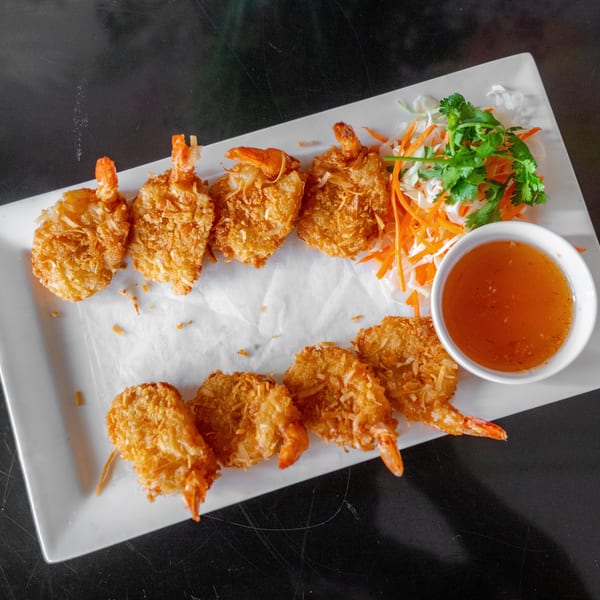 A white rectangular plate holds eight golden-brown coconut shrimp, a side of shredded vegetables, and a dipping sauce.