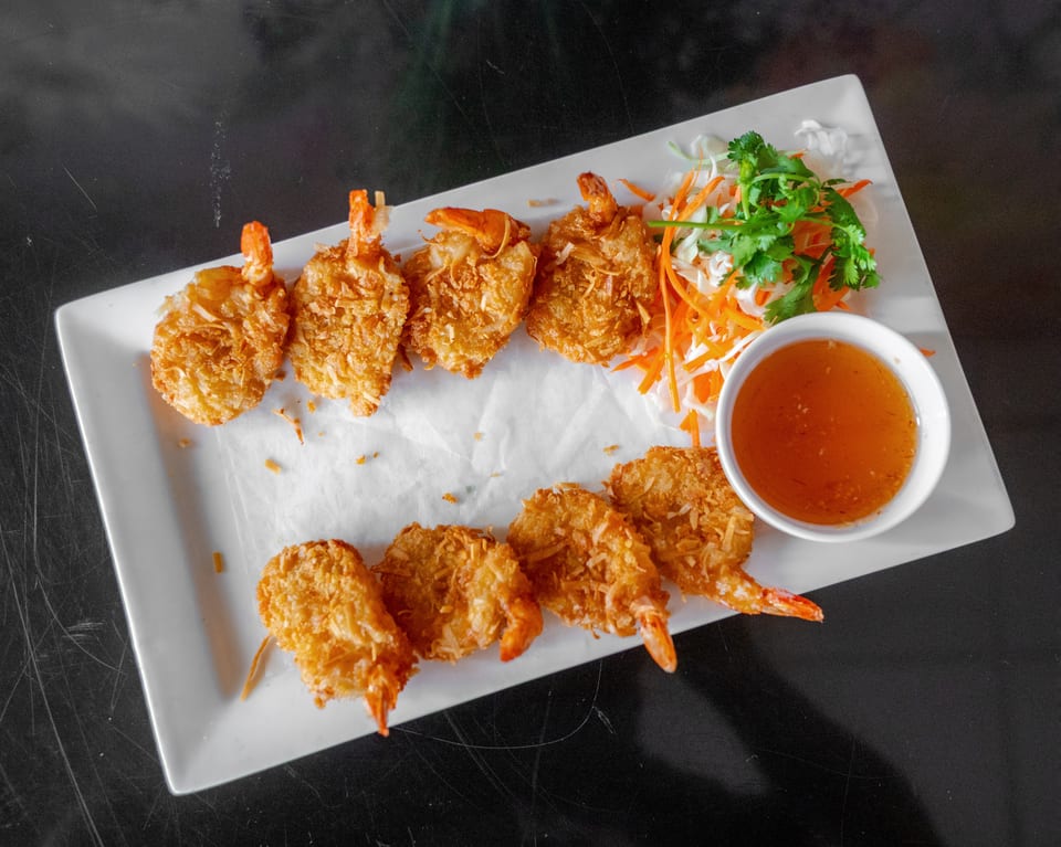 A white rectangular plate holds eight golden-brown coconut shrimp, a side of shredded vegetables, and a dipping sauce.