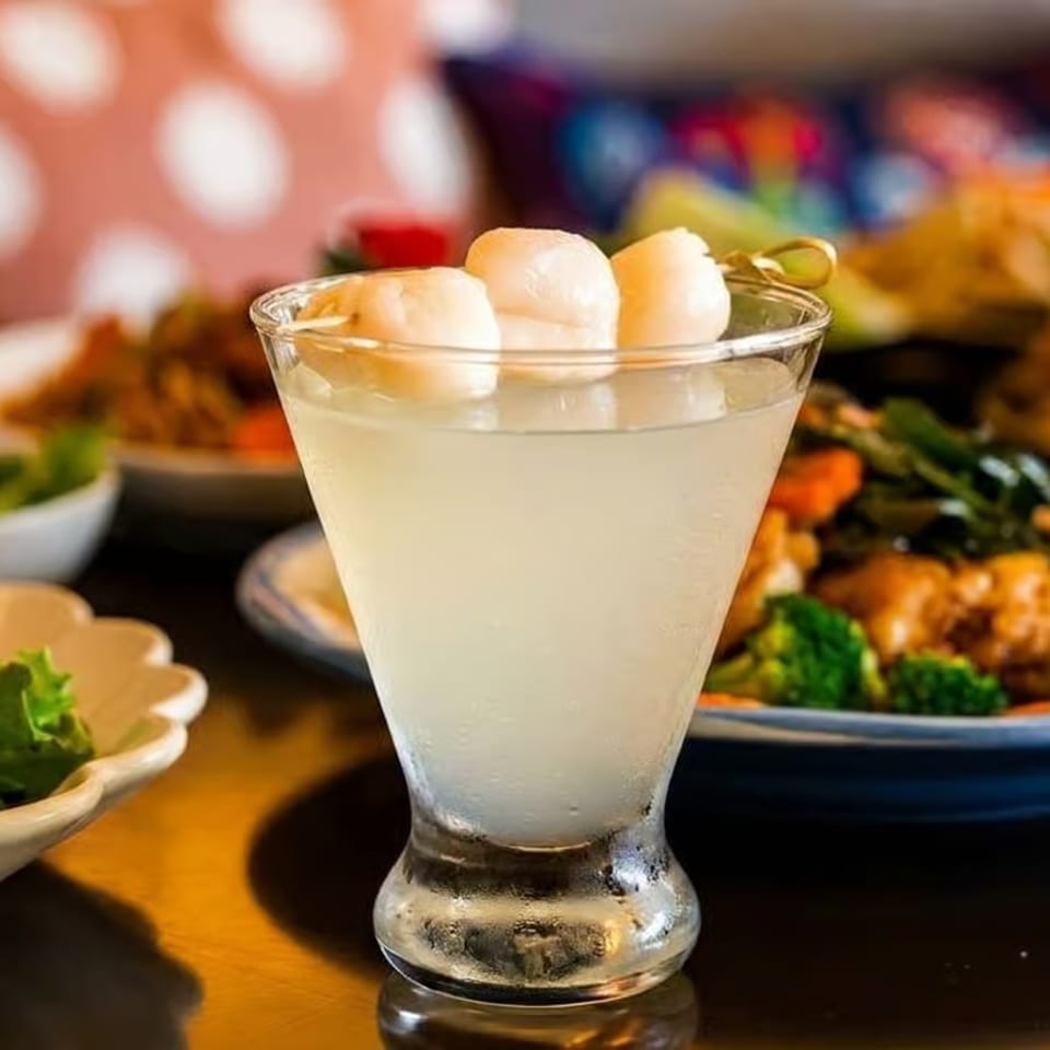Lychee martini with three lychees on a skewer, surrounded by various Thai dishes and drinks.