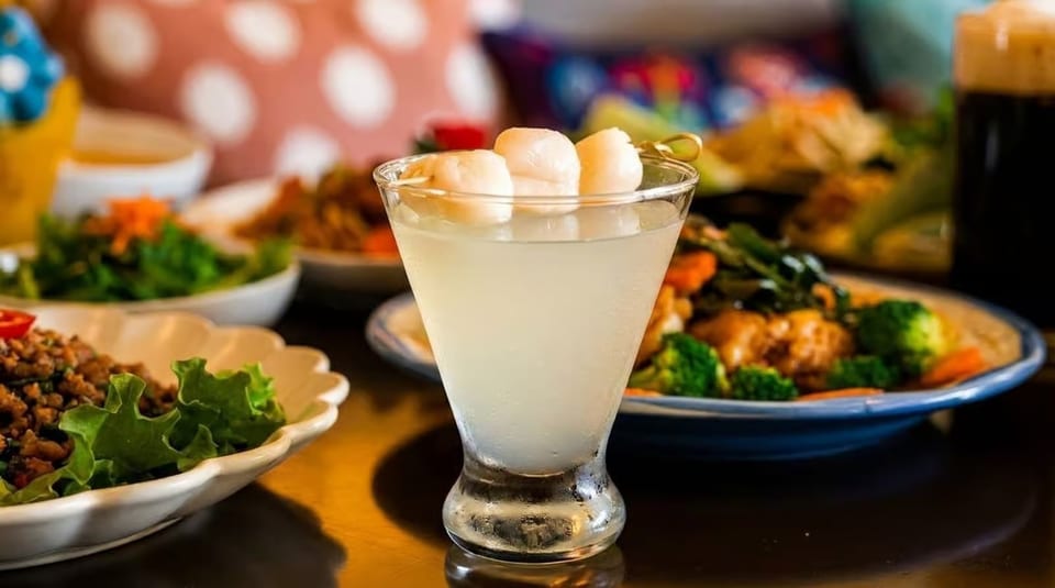 Lychee martini with three lychees on a skewer, surrounded by various Thai dishes and drinks.