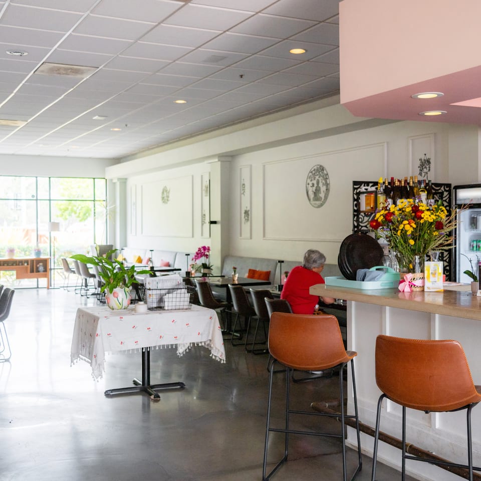 Bright, spacious restaurant interior with a bar, dining tables, and a large window overlooking trees.