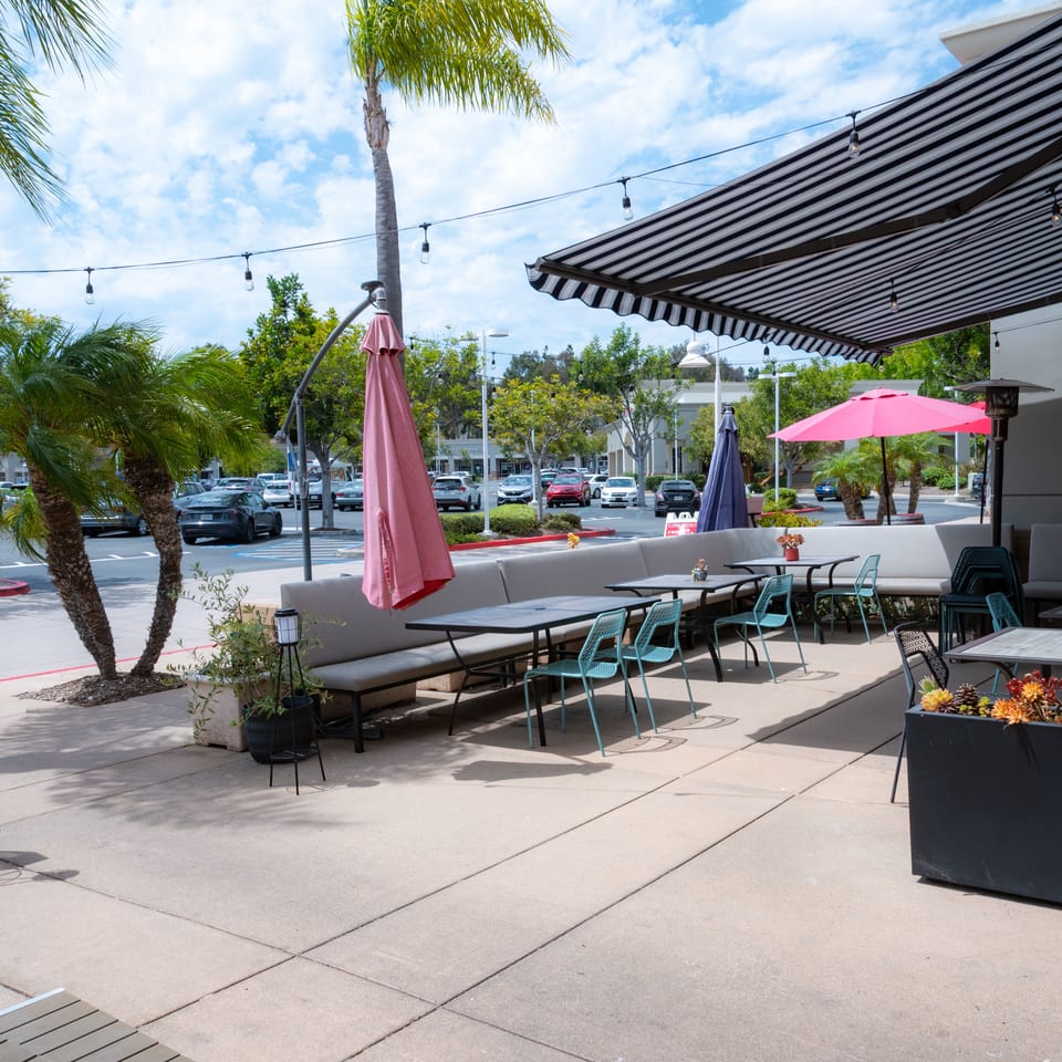 Outdoor restaurant patio with tables, chairs, umbrellas, and palm trees under a striped awning.