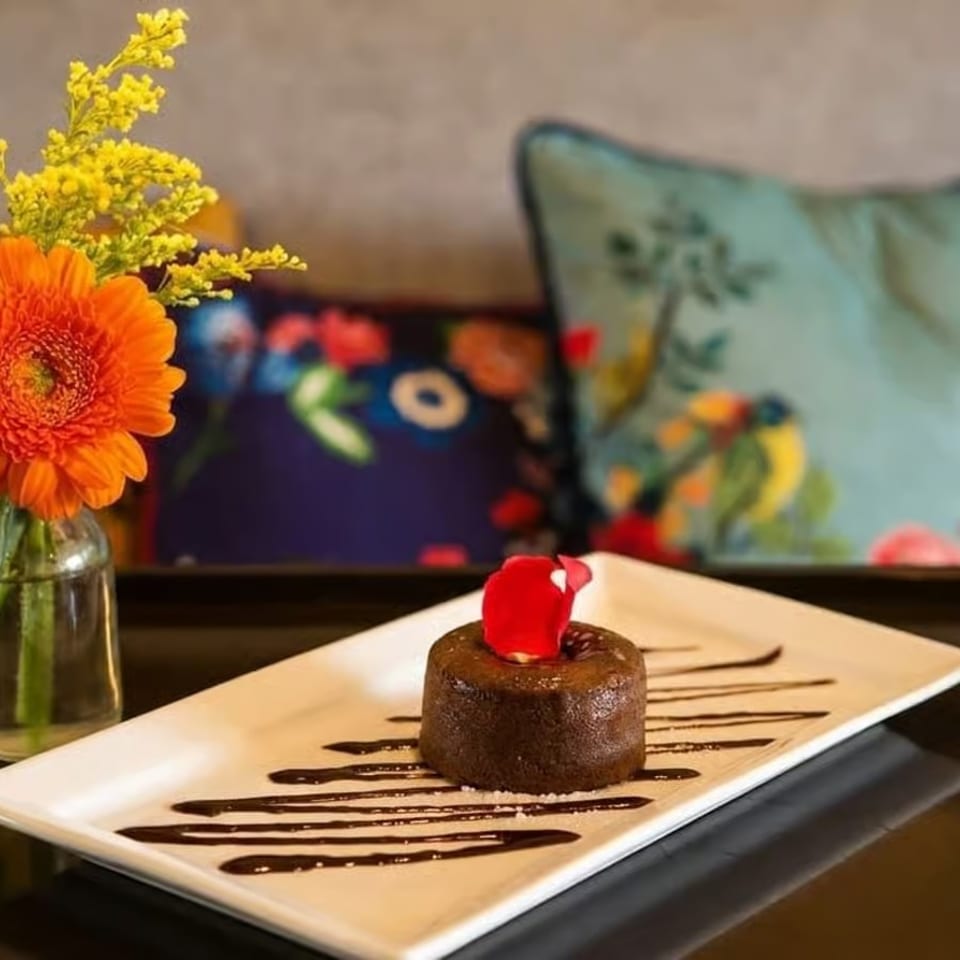 Chocolate lava cake with a red petal on a white plate, next to a vase of orange and yellow flowers.