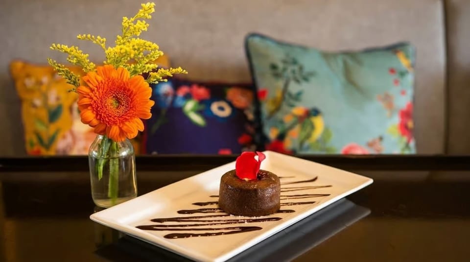 Chocolate lava cake with a red petal on a white plate, next to a vase of orange and yellow flowers.
