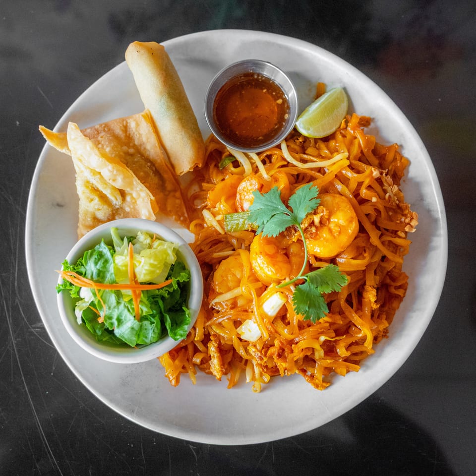 Pad Thai with shrimp, egg roll, fried wontons, side salad, and dipping sauce on a white plate.