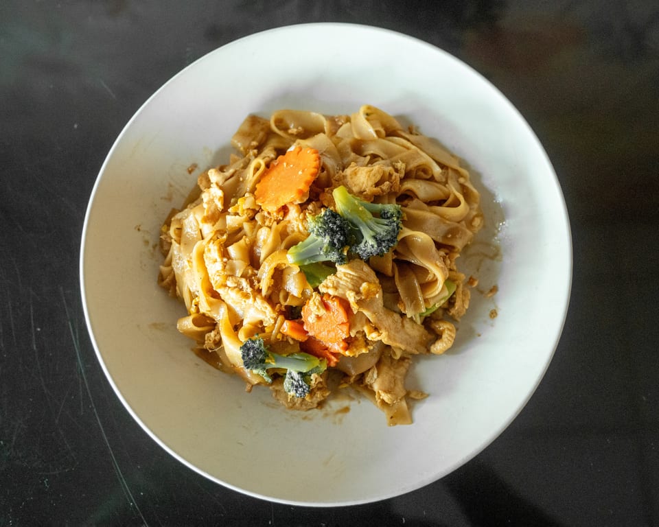 Pad See Ew with chicken, broccoli, and carrots in a white bowl on a dark surface.
