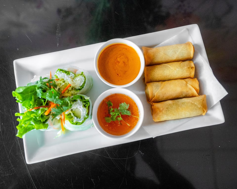 A white rectangular plate holds fresh spring rolls, fried egg rolls, and two dipping sauces.