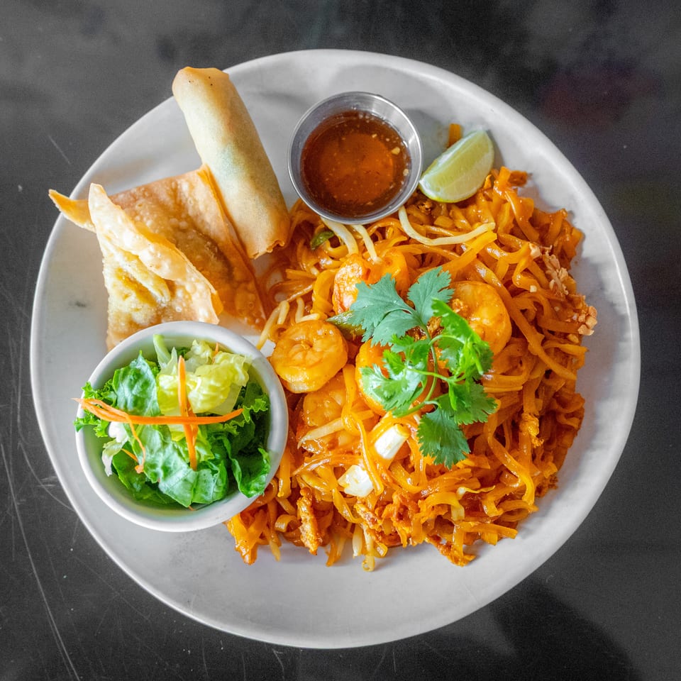 Shrimp Pad Thai with a side salad, spring roll, crispy wontons, and dipping sauce on a white plate.
