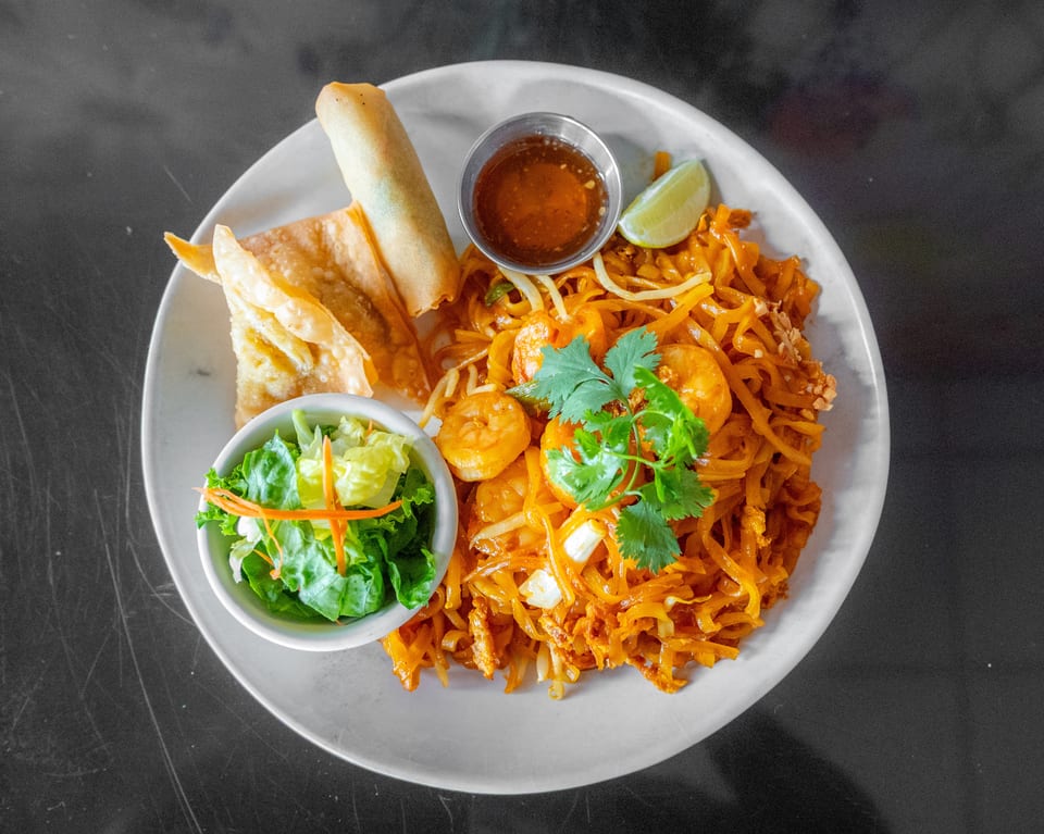 Shrimp Pad Thai with a side salad, spring roll, crispy wontons, and dipping sauce on a white plate.