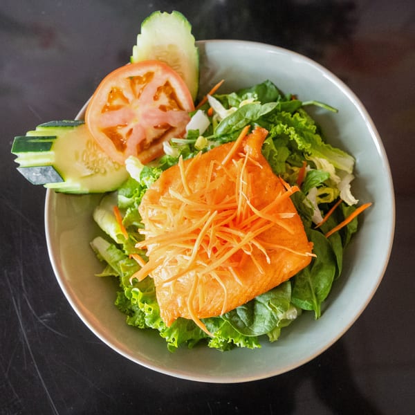 A fresh salad with greens, sliced cucumbers, tomatoes, and a salmon fillet topped with shredded carrots.