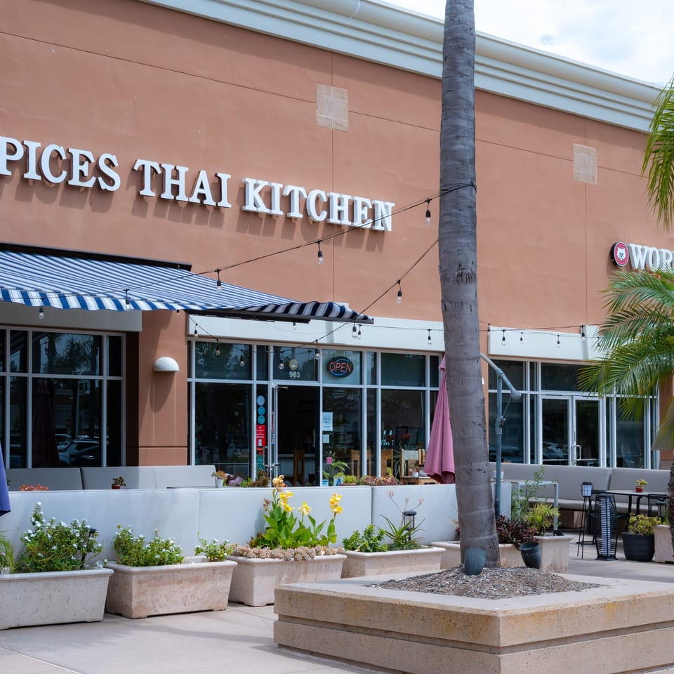 Spices Thai Kitchen restaurant exterior with outdoor seating, striped awning, and palm trees.