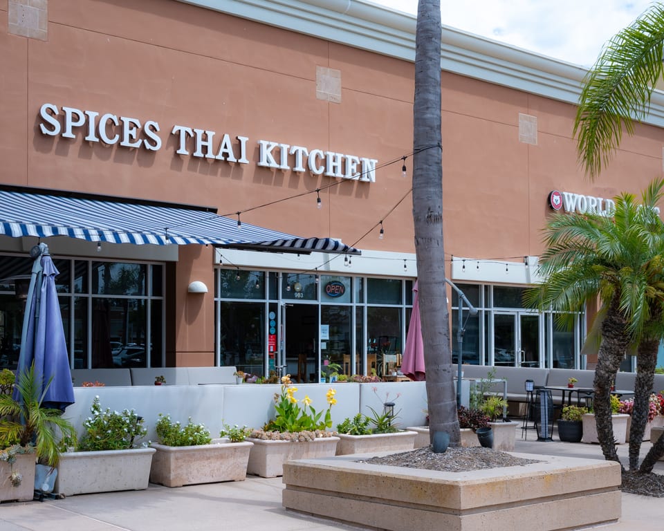 Spices Thai Kitchen restaurant exterior with outdoor seating, striped awning, and palm trees.